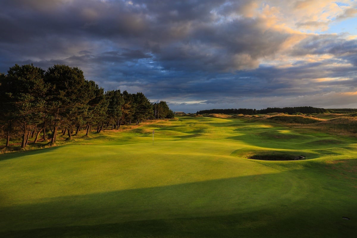 Golf course fairway lined with trees under a dramatic cloudy sky at sunset